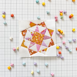 Może przedstawiać: Blok patchworkowej kołdry z wzorem gwiazdy w odcieniach różowego, żółtego i brązowego. Blok jest otoczony małymi origami gwiazdami w różnych kolorach, w tym różowym, żółtym, niebieskim i pomarańczowym.