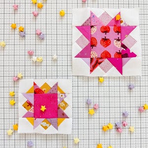 Może przedstawiać: Dwa bloki patchworkowej kołdry w kolorze różowym i białym z wzorem gwiazdy. Bloki znajdują się na białej powierzchni w kratkę z rozrzuconymi wokół małymi origami gwiazdami.