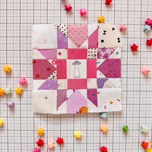 Może przedstawiać: Kwadratowy blok patchworkowy z różowych, fioletowych i białych tkanin. Na środku znajduje się ilustracja grzyba. Kolorowe gwiazdki origami otaczają blok na tle kratki.