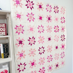 Może przedstawiać: Różowa i biała kołdra z wzorem gwiazd. Kołdra składa się z wielu różnych kwadratów tkaniny, każdy z innym wzorem gwiazdy. Kołdra wisi na białej ścianie.