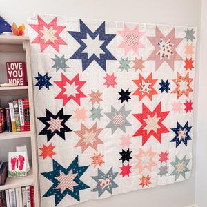 May include: A large, handmade quilt featuring a star pattern in various colors, including red, blue, pink, and peach, against a white background. The quilt is hanging on a wall. The text "LOVE YOU MORE" is visible on a shelf.