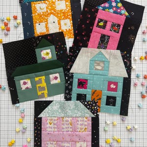 Peut inclure: Cinq blocs de courtepointe en tissu représentant des maisons aux couleurs et motifs variés. Les blocs comprennent une maison jaune, une rose, une verte, une bleu clair et une rose pâle, toutes assemblées à partir de pièces de tissu en coton.