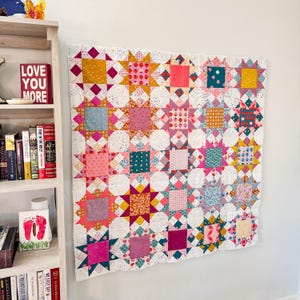 May include: A white quilt with a patchwork design featuring pink, red, yellow, blue, and green stars. The quilt is hanging on a white wall.