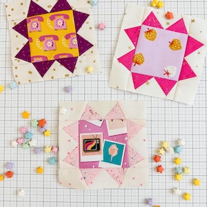 Puede incluir: Tres bloques de edredón coloridos en forma de estrella con varios diseños. Un bloque presenta un fondo amarillo con teléfonos rosas, otro tiene fresas y el último imágenes retro. Estrellas de origami dispersas en varios colores rodean los bloques.