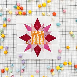 Może przedstawiać: Biały kwadratowy kawałek materiału z wzorem rozbłysku gwiazdy w kolorze różowym i czerwonym. Materiał jest otoczony małymi gwiazdami origami w różnych kolorach, w tym różowym, żółtym, niebieskim i białym.