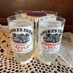 Może przedstawiać: Przezroczysty plastikowy uchwyt na napoje z czterema szklankami. Każda szklanka ma logo "Boone's Farm" i tekst dla "Apple Wine" i "Strawberry Hill". Uchwyt jest biały i stoi na białym koronkowej obrusie.