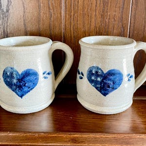Pode incluir: Duas canecas de cerâmica creme, cada uma com um grande coração azul. Cada caneca tem uma alça e é decorada com pequenos detalhes florais azuis. As canecas são exibidas em uma superfície de madeira.