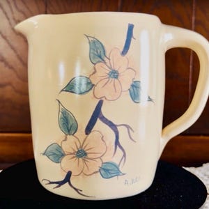 May include: A cream-colored ceramic pitcher featuring a hand-painted design of two light orange flowers with green leaves on dark blue branches. The pitcher has a curved handle and a pouring spout, with the initials A.D. written near the base.
