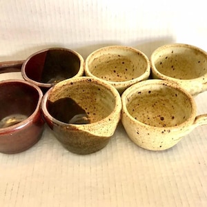 May include: A collection of six ceramic cups arranged in two rows. The front row features a solid dark brown cup, a speckled brown and cream cup, and a speckled cream cup with a handle. The back row includes a dark brown cup with a handle, and two speckled cream cups.
