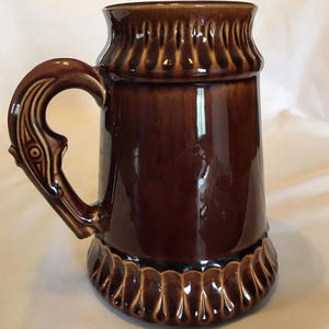 May include: A brown ceramic beer stein with a decorative handle and base. The stein has a glossy finish and a detailed, textured design around the top and bottom. The handle features an ornate, stylized design.