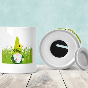 Könnte beinhalten: Weiße Keramik-Sparbüchse mit einem grünen Zwerg, der einen grünen Hut mit einer gelben Blume und grünen Blättern trägt. Der Zwerg ist von grünem Gras umgeben. Die Sparbüchse hat einen Schlitz für Münzen oben.