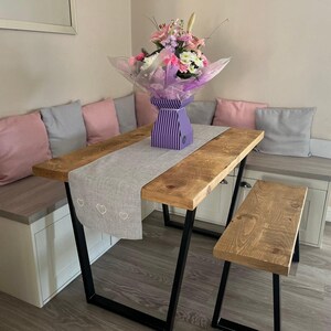 May include: A wooden dining table with black metal legs and a matching bench. The table has a grey linen runner with white hearts. A bouquet of pink and purple flowers in a purple and white striped vase sits on the table.