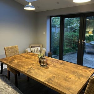 May include: A large, rectangular wooden dining table with a warm, natural finish. The table is surrounded by woven chairs and a bench. Two pendant lights hang above, illuminating the space. A decorative lantern and flowers sit on the table.