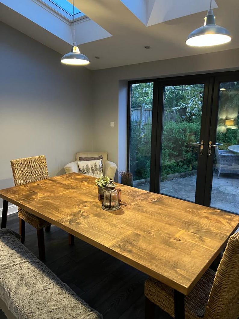 May include: A large, rectangular wooden dining table with a warm, natural finish. The table is surrounded by woven chairs and a bench. Two pendant lights hang above, illuminating the space. A decorative lantern and flowers sit on the table.