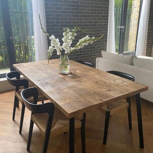 May include: A rectangular wooden dining table with black legs and four chairs. The chairs have black frames and woven seats. A glass vase with white flowers sits on the table. The room has a brick wall and large windows.