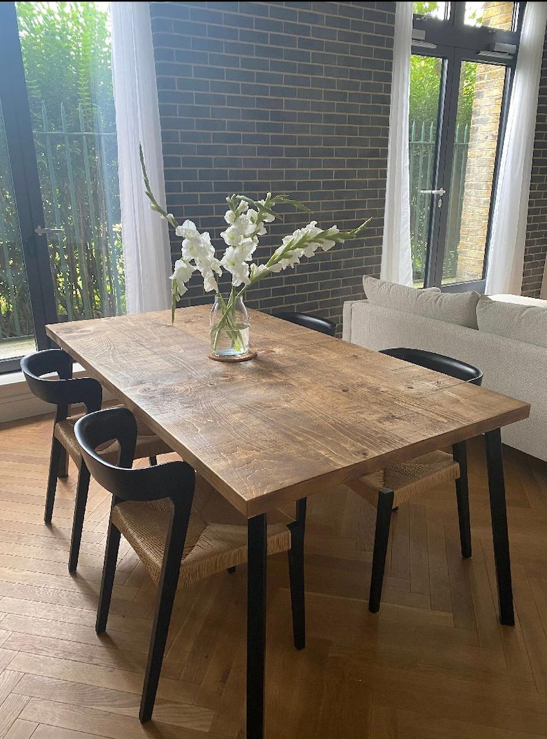 May include: A rectangular wooden dining table with black legs and four chairs. The chairs have black frames and woven seats. A glass vase with white flowers sits on the table. The room has a brick wall and large windows.