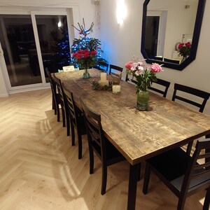 May include: A long, rustic wooden dining table with black legs, set with flowers and candles. The table is surrounded by black chairs. The room has light-colored wood flooring and a large window.