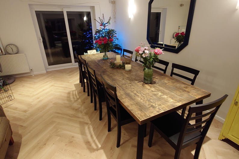 May include: A long, rustic wooden dining table with black legs, set with flowers and candles. The table is surrounded by black chairs. The room has light-colored wood flooring and a large window.