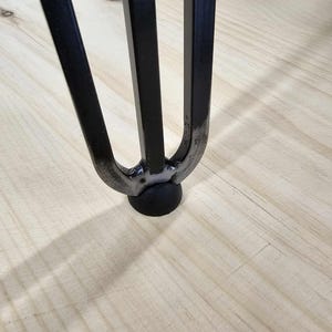 May include: Close-up of a black metal hairpin leg with a round black foot pad. The leg is resting on a light wood surface.