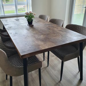 May include: A rectangular wooden dining table with a dark brown finish, supported by black metal legs. Six upholstered chairs with diamond-patterned backs surround the table. A small potted plant with pink flowers sits on the table's surface. The table is approximately 72 inches long.