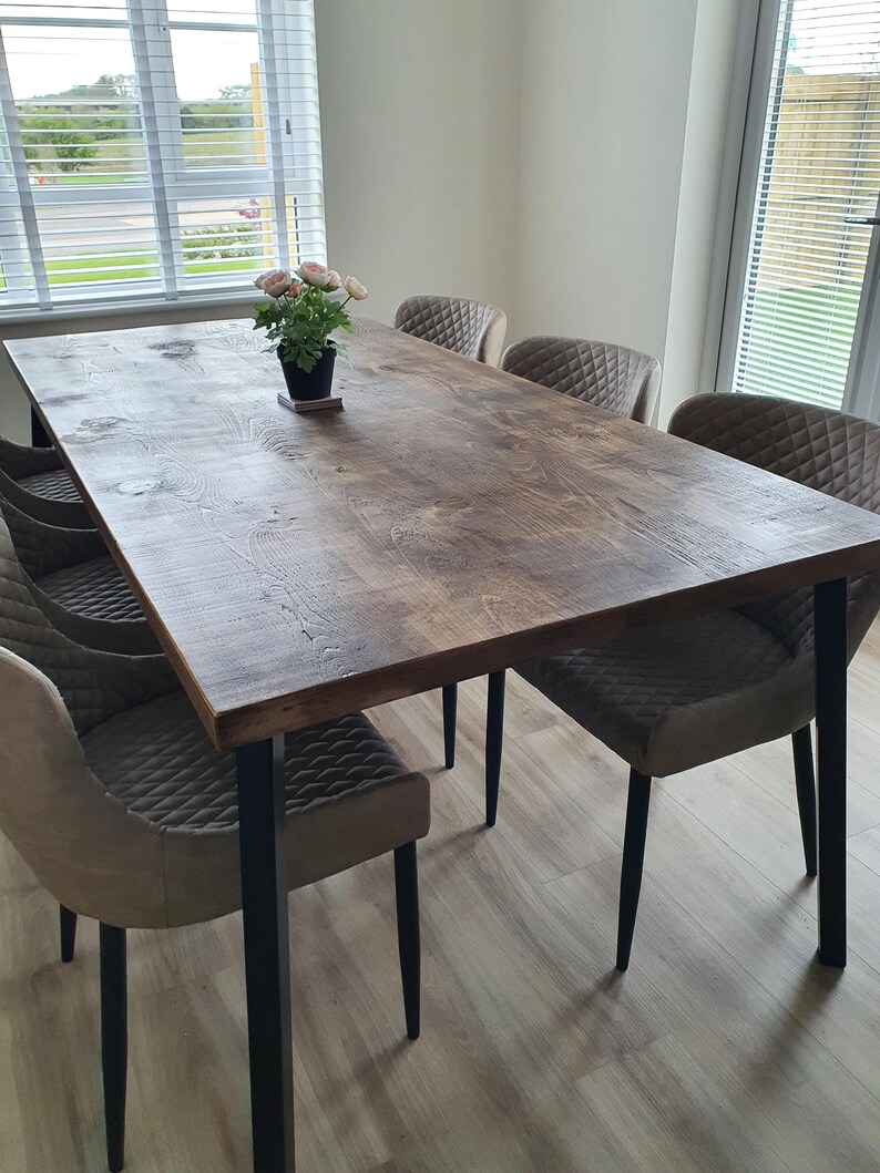 May include: A rectangular wooden dining table with a dark brown finish, supported by black metal legs. Six upholstered chairs with diamond-patterned backs surround the table. A small potted plant with pink flowers sits on the table's surface. The table is approximately 72 inches long.