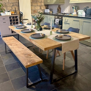May include: A rustic wooden dining table with a matching bench, set for a meal. The table has a light brown wood top and black metal legs. The table is set with plates, placemats, and candles. The kitchen has green cabinets and a black tiled floor.