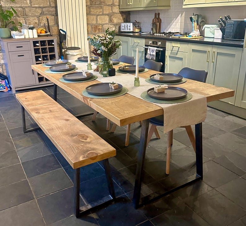 May include: A rustic wooden dining table with a matching bench, set for a meal. The table has a light brown wood top and black metal legs. The table is set with plates, placemats, and candles. The kitchen has green cabinets and a black tiled floor.