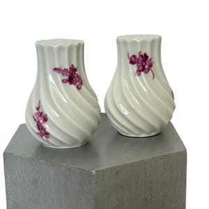 May include: A pair of white ceramic salt and pepper shakers with a spiral design. Each shaker features a purple floral pattern. The shakers are displayed on a gray surface.