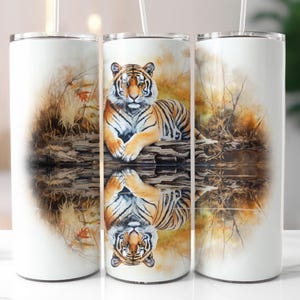May include: A white stainless steel tumbler with a watercolor design of a tiger sitting in a stream. The tiger is looking directly at the viewer. The design is reflected in the water.