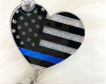 May include: A heart-shaped badge holder with a thin blue line American flag design. The holder is made of clear resin with glitter embedded in the design. The holder has a silver clip and a small round charm with a thin blue line American flag design.
