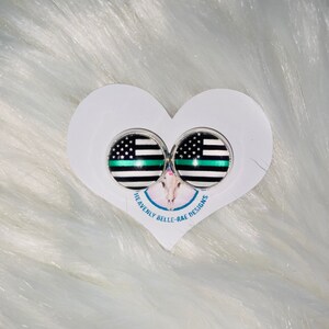 May include: A pair of silver stud earrings with a black and white thin blue line flag design. The earrings are displayed on a white heart-shaped card with the text "Heavenly Belle-Rae Designs".