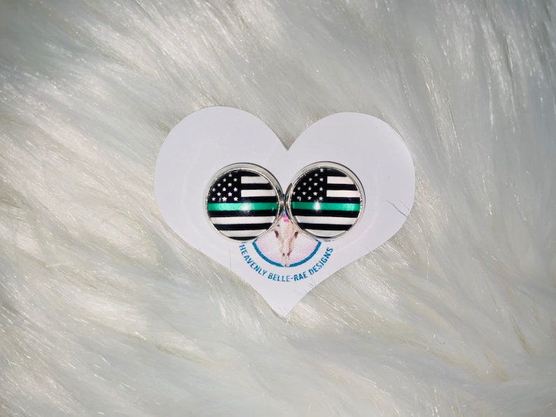 May include: A pair of silver stud earrings with a black and white thin blue line flag design. The earrings are displayed on a white heart-shaped card with the text "Heavenly Belle-Rae Designs".