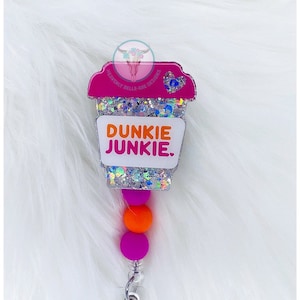 May include: A pink and white glitter coffee cup charm with the text "Dunkie Junkie" on a white background. The charm is attached to a clear retractable badge holder with three colorful beads.
