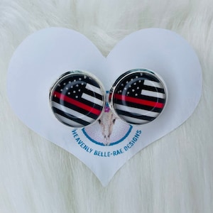 May include: A pair of silver-toned stud earrings with a red, white, and black American flag design with a thin red stripe across the middle. The earrings are on a white heart-shaped card with the text "Heavenly Belle-Rae Designs".