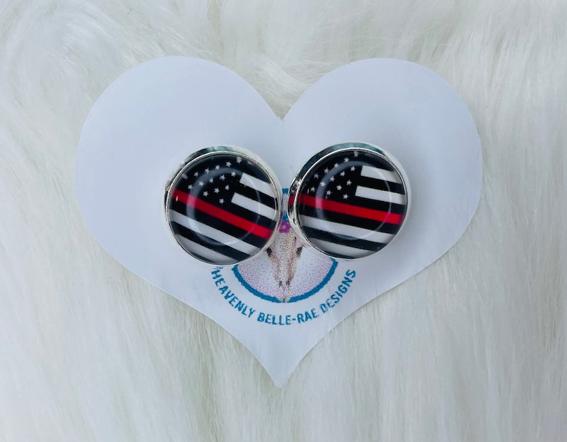 May include: A pair of silver-toned stud earrings with a red, white, and black American flag design with a thin red stripe across the middle. The earrings are on a white heart-shaped card with the text "Heavenly Belle-Rae Designs".