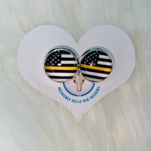 May include: A pair of silver stud earrings with a black and white American flag design with a yellow stripe. The earrings are on a white heart-shaped card with the text "Heavenly Belle-Rae Designs".