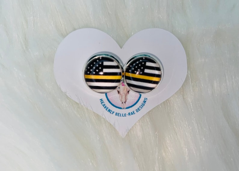 May include: A pair of silver stud earrings with a black and white American flag design with a yellow stripe. The earrings are on a white heart-shaped card with the text "Heavenly Belle-Rae Designs".