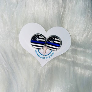 May include: A pair of silver stud earrings with a thin blue line American flag design. The earrings are displayed on a white heart-shaped card with the text "Heavenly Belle-Rae Designs".