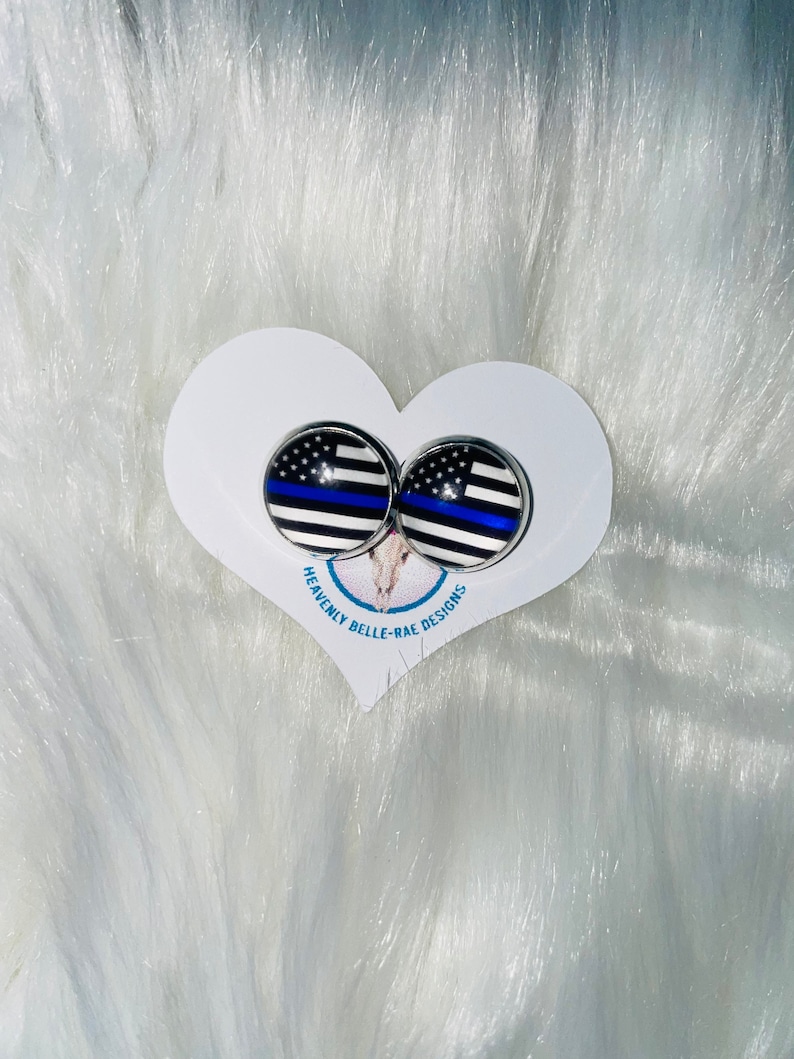 May include: A pair of silver stud earrings with a thin blue line American flag design. The earrings are displayed on a white heart-shaped card with the text "Heavenly Belle-Rae Designs".