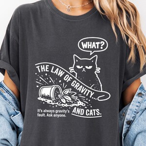 May include: Dark gray t-shirt with a humorous graphic. The design features a cat with an angry expression and the text "WHAT?" in a speech bubble. Below, it reads "THE LAW OF GRAVITY... AND CATS. It's always gravity's fault. Ask anyone."