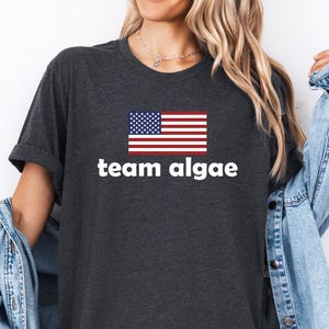 Peut inclure: Un t-shirt gris anthracite foncé arborant un graphique du drapeau des États-Unis centré au-dessus du texte blanc en minuscules indiquant team algae. Le t-shirt a des manches courtes et un col rond, porté avec une veste en jean bleu clair par-dessus.
