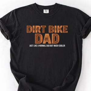 May include: Black t-shirt featuring the text "DIRT BIKE DAD" in orange block letters, with a tyre track design in the word "DAD". The phrase "JUST LIKE A NORMAL DAD BUT MUCH COOLER" is printed below in white.