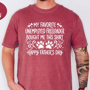 May include: Crimson-colored t-shirt with white text that reads "MY FAVORITE UNEMPLOYED FREELOADER BOUGHT ME THIS SHIRT" above two paw prints and the words "Happy Father's Day". The shirt is short-sleeved and worn by a person.