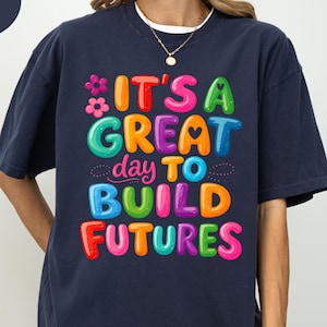 Pode incluir: Camiseta azul marinho com a frase "IT'S A GREAT day TO BUILD FUTURES" em letras coloridas e em bolhas. O design inclui uma flor e um cora&ccedil;&atilde;o. A camiseta &eacute; de estilo confort&aacute;vel e casual.