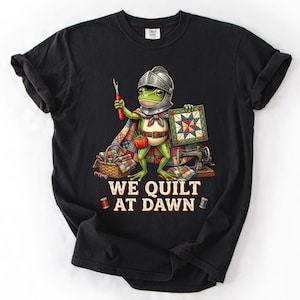 Peut inclure: Un t-shirt noir avec une illustration représentant une grenouille verte portant un casque en métal et une cape, tenant un découd-vite et un bloc de patchwork. À côté se trouvent une machine à coudre et un panier. Le texte indique WE QUILT AT DAWN.