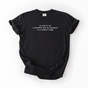 Może przedstawiać: Czarny t-shirt z białym napisem: "be nice to me I probably lost an argument to a toddler today." Koszulka z kr&oacute;tkim rękawem wykonana jest z miękkiego materiału. Koszulka jest prezentowana na białym tle.