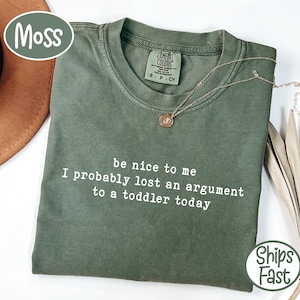 Może przedstawiać: Zielona koszulka w kolorze mchu z białym napisem "be nice to me I probably lost an argument to a toddler today". Koszulka ma okrągły dekolt i małą metkę na kołnierzu. Złoty naszyjnik z wisiorkiem jest zarzucony na koszulkę.