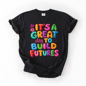 Pode incluir: Camiseta preta com a frase "IT'S A GREAT day TO BUILD FUTURES" em letras coloridas e em bolhas. O design inclui flores e pontos. A camiseta &eacute; feita de um material macio.