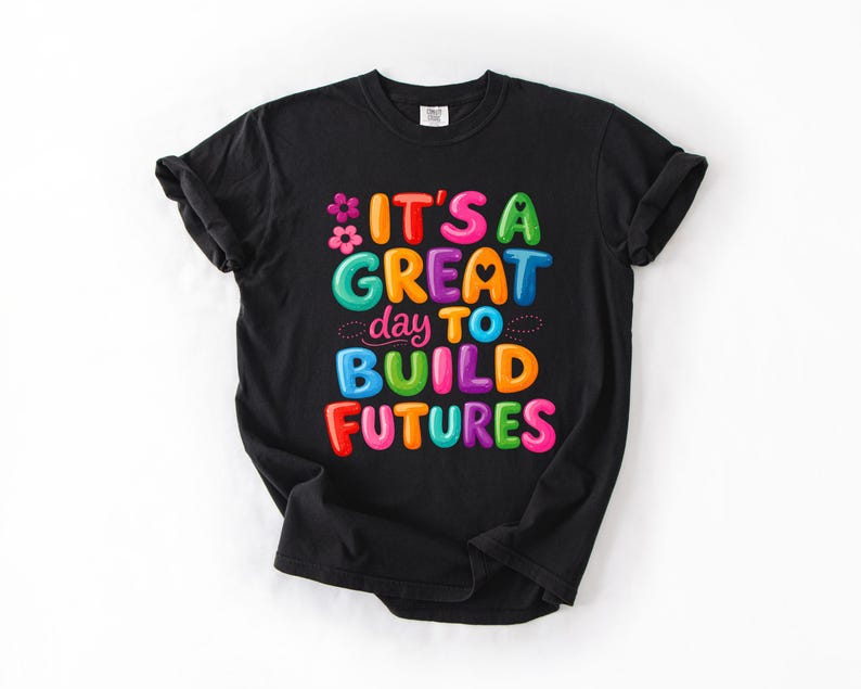 Pode incluir: Camiseta preta com a frase "IT'S A GREAT day TO BUILD FUTURES" em letras coloridas e em bolhas. O design inclui flores e pontos. A camiseta &eacute; feita de um material macio.