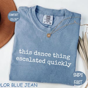 以下が含まれることがあります： 「this dance thing escalated quickly」の白文字がプリントされた、ブルーデニムカラーのTシャツ。ペンダント付きのゴールドネックレスがシャツにかけられています。シャツは白い面に折りたたまれています。